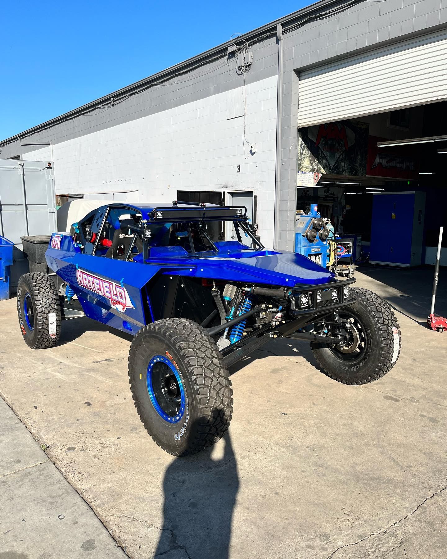 Blue off-road race buggy parked outside a garage in bright sunlight.