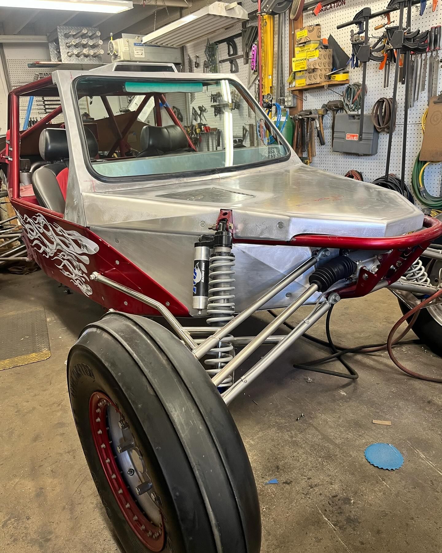 Red and silver off-road buggy in a workshop, front-left view with exposed suspension and tools on the wall.