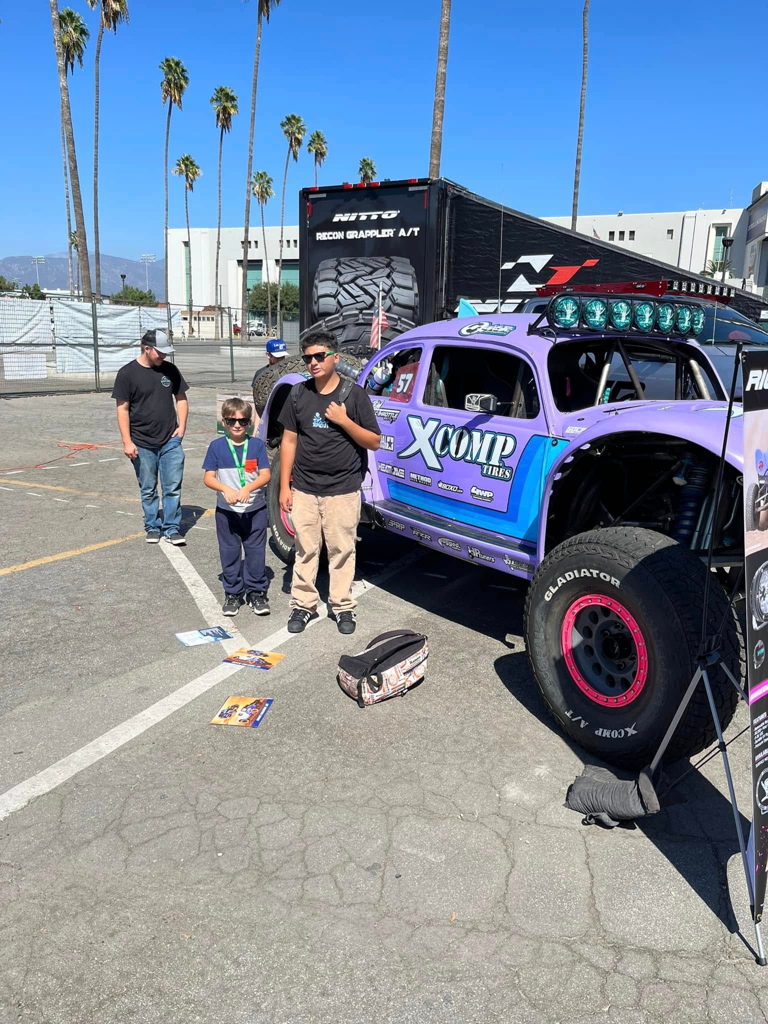 People posing beside a purple off-road truck at an outdoor event with palm trees and tents.