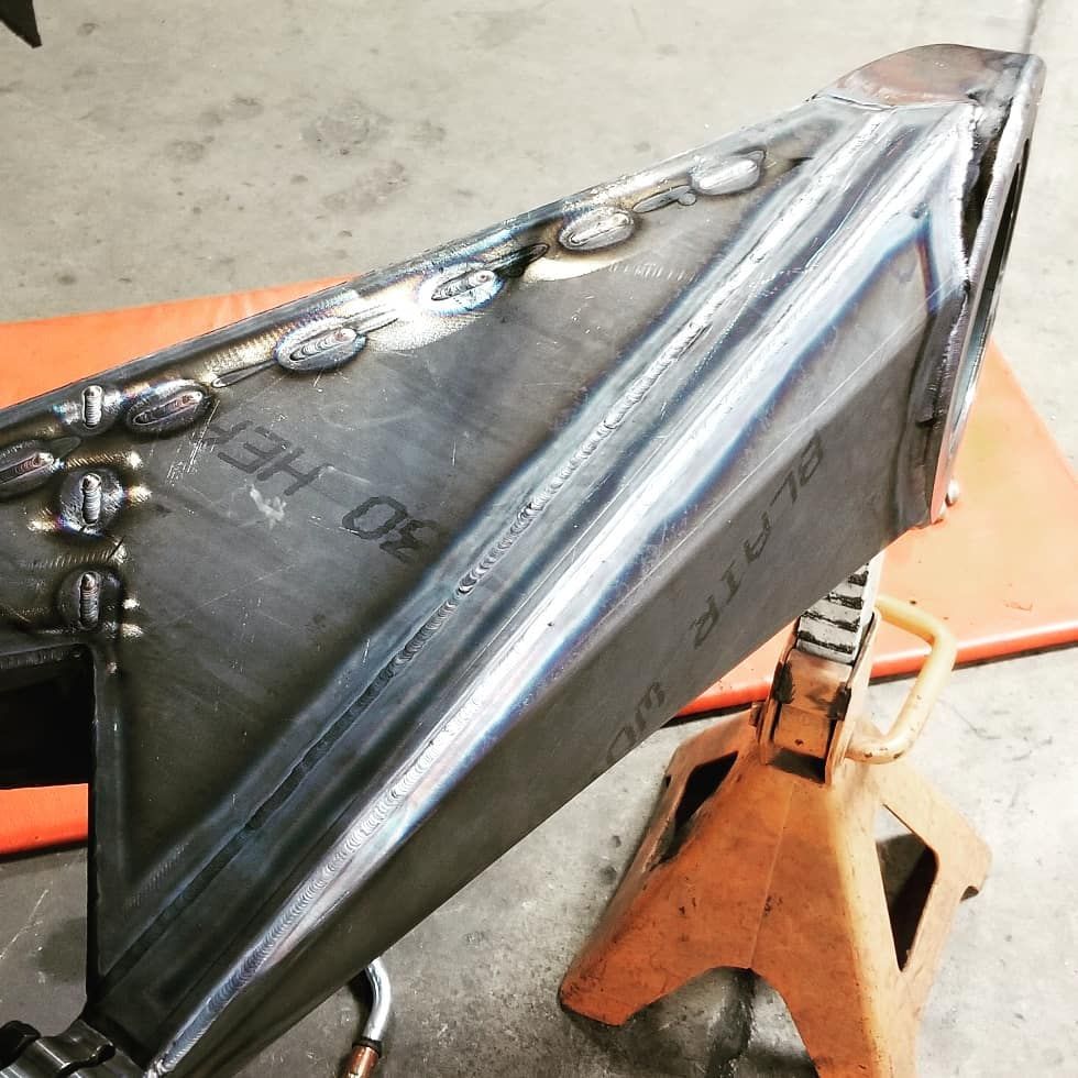 Close-up of a welded black metal part clamped in a vise, with orange tools and surface rust visible.
