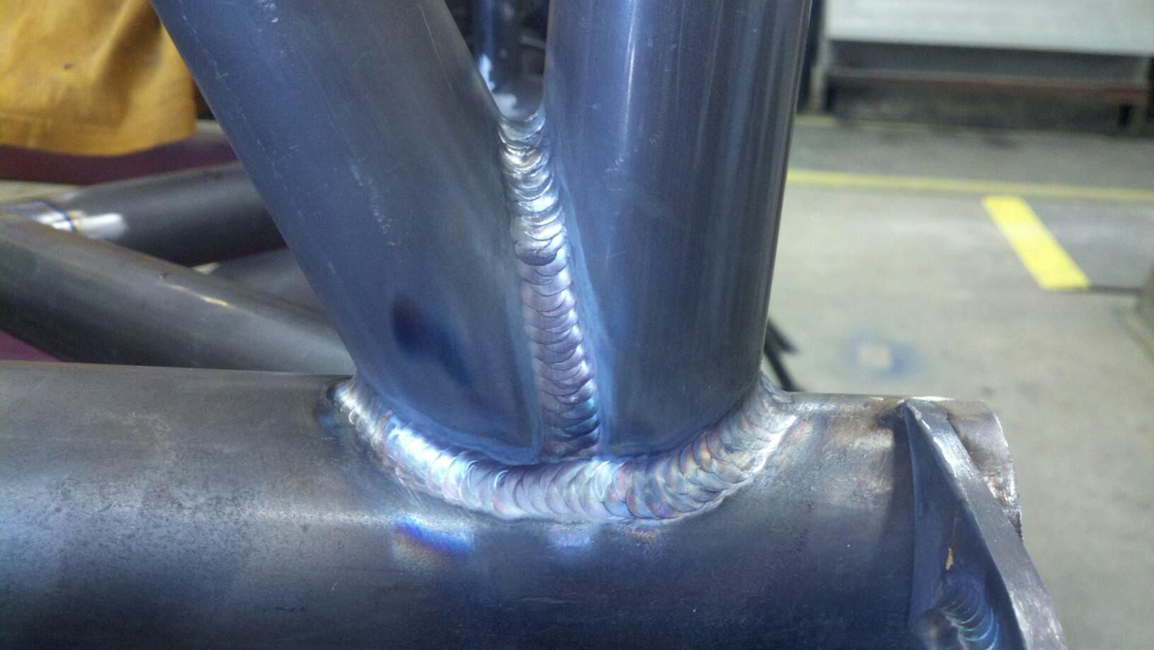 Close-up of a welded metal pipe joint with bright blue-gray weld bead on a workshop floor