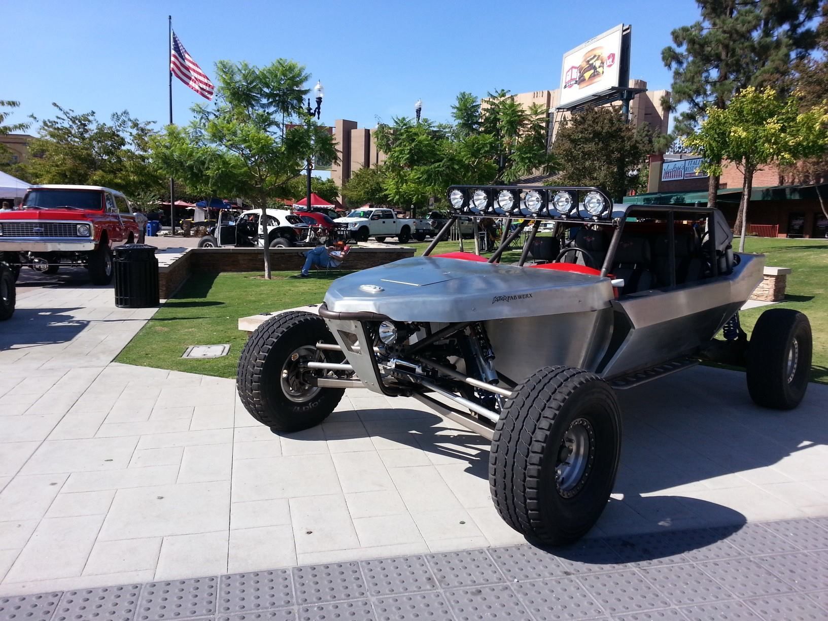 Off-road dune buggy parked on a sunny sidewalk near trees and a U.S. flag