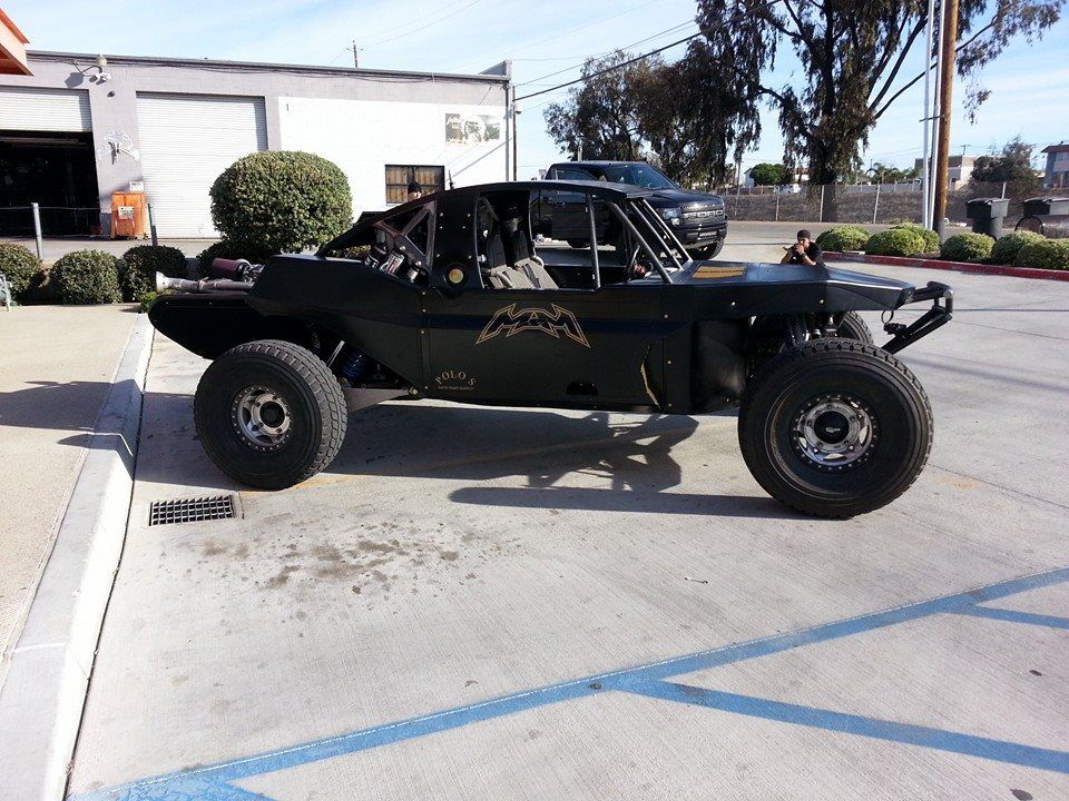 Black off-road buggy with large tires parked in a lot near a building and palm trees.