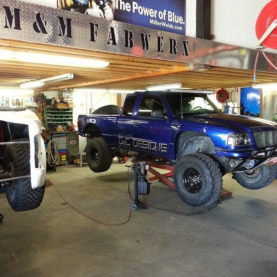 Blue lifted pickup truck in an auto shop with another truck partially visible nearby