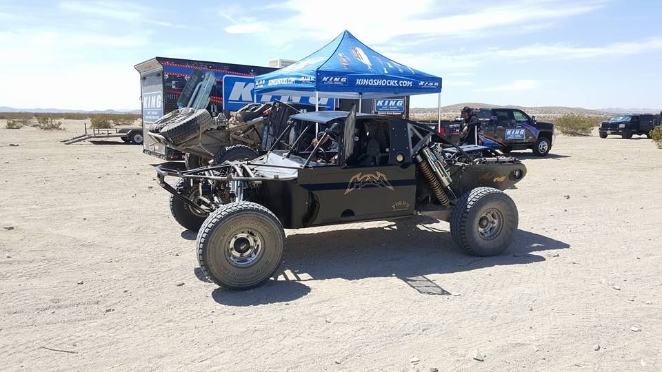 Off-road buggy parked in a desert, with a blue canopy tent and other vehicles in the background.