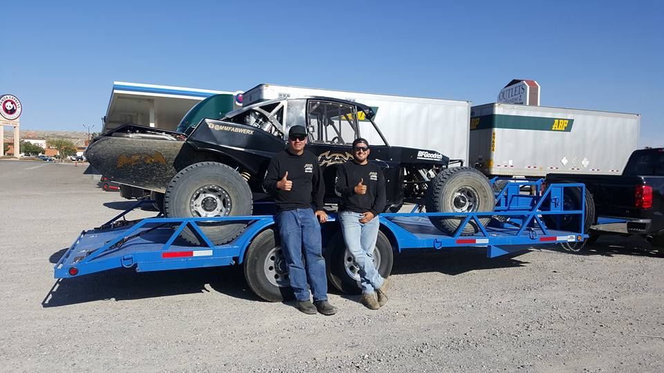 Two men lean on a blue car trailer in front of a black off-road buggy in a parking lot