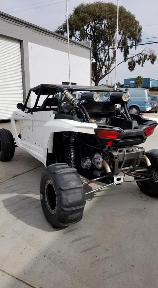 White off-road dune buggy parked outdoors near a building, with exposed engine and oversized rear tires