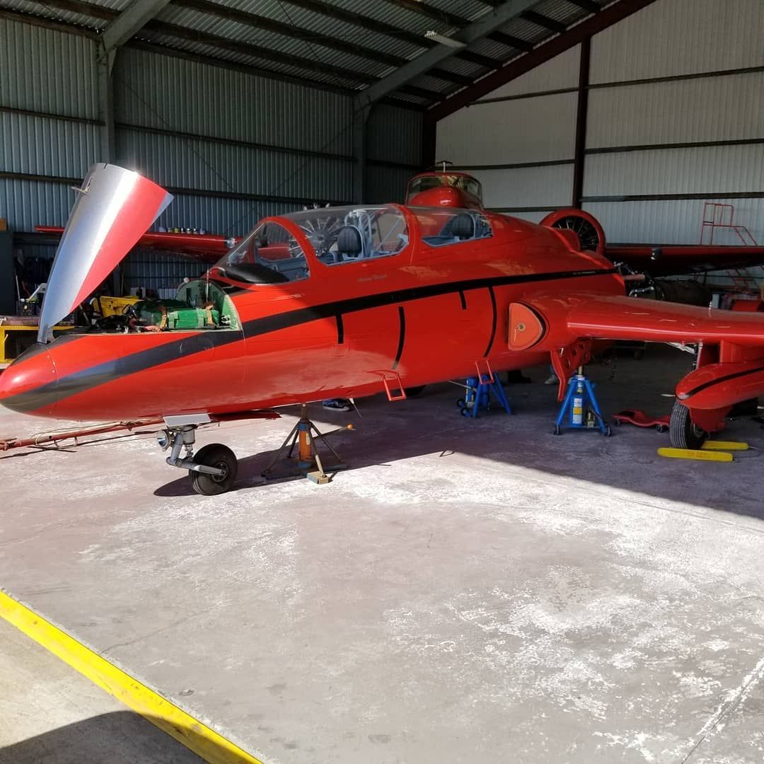 Red aircraft in a hangar, with a person seated in the cockpit and maintenance gear nearby