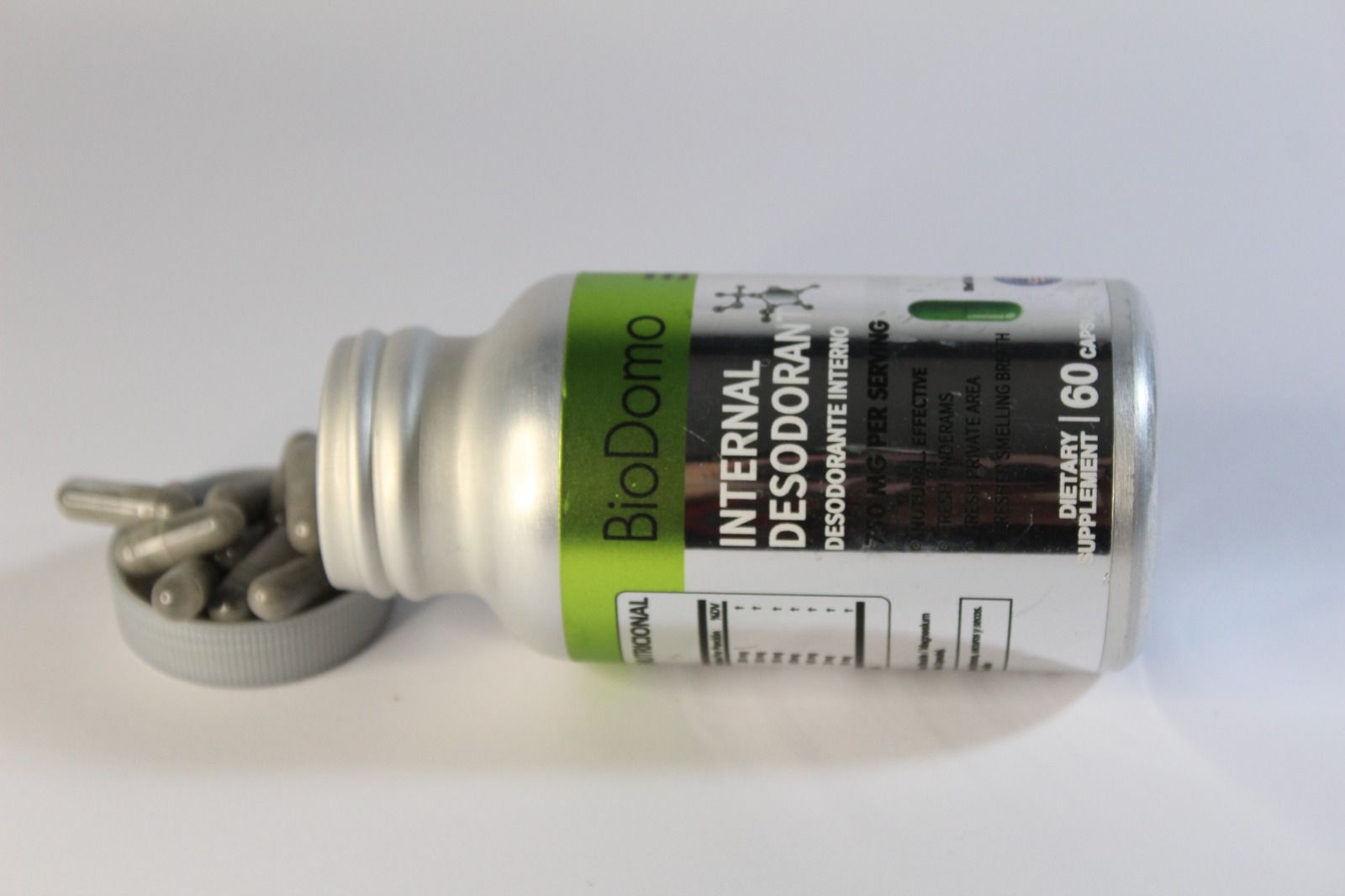 Botella plateada de cápsulas de "Desodorante Interno" derramada sobre blanco.