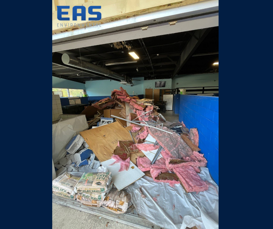 Workers in safety gear on a roof removing material with abatement equipment; urban setting with buildings in background.