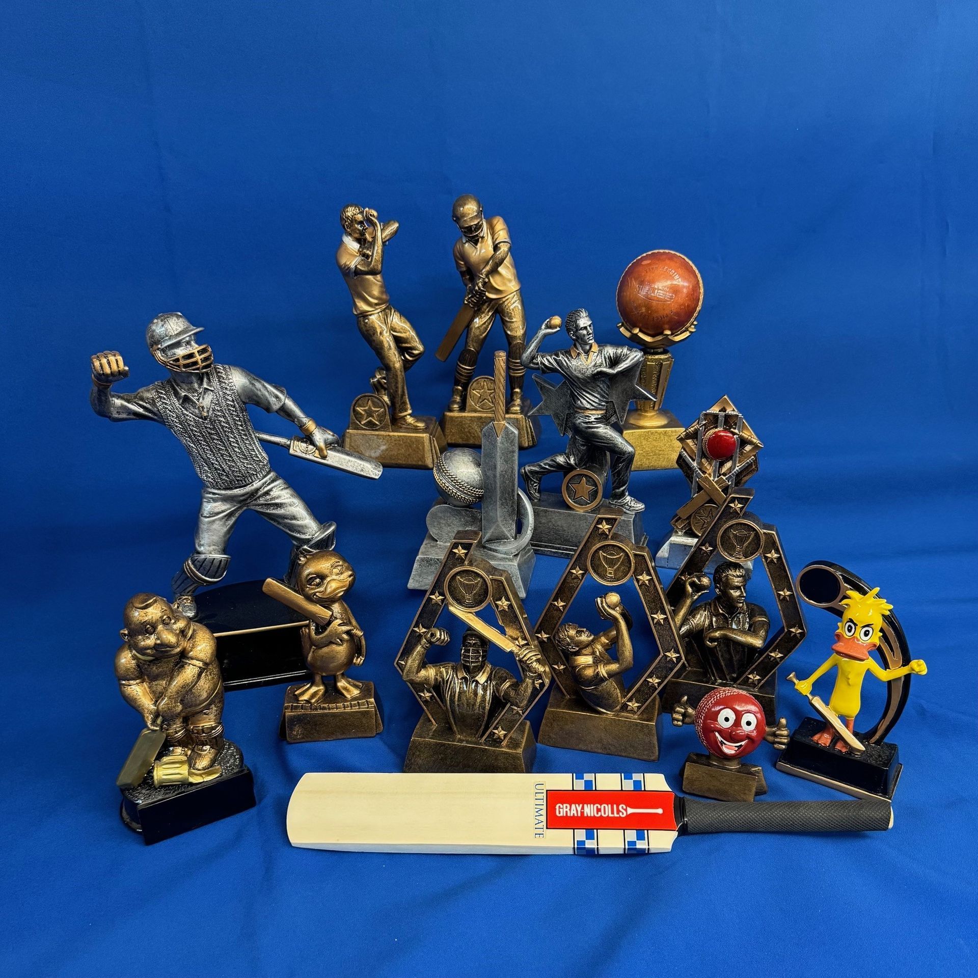 Cricket trophies and a bat on a blue surface. Bronze and silver figures in action — NQ Plaques & Trophies In Mount Isa, QLD