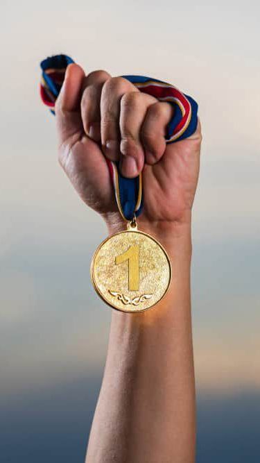 An arm in front of a blurry background is holding a first place medal, displaying it proudly.