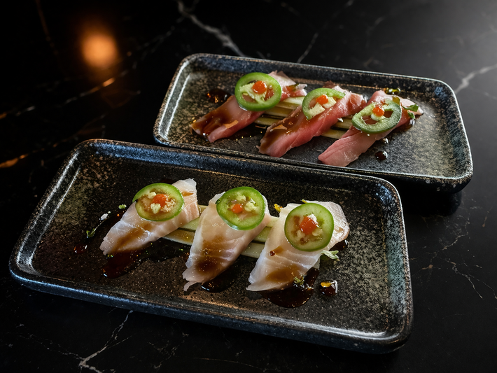 Two dark, speckled rectangular plates holding slices of sashimi topped with jalapeño and sauce on a black marble table.