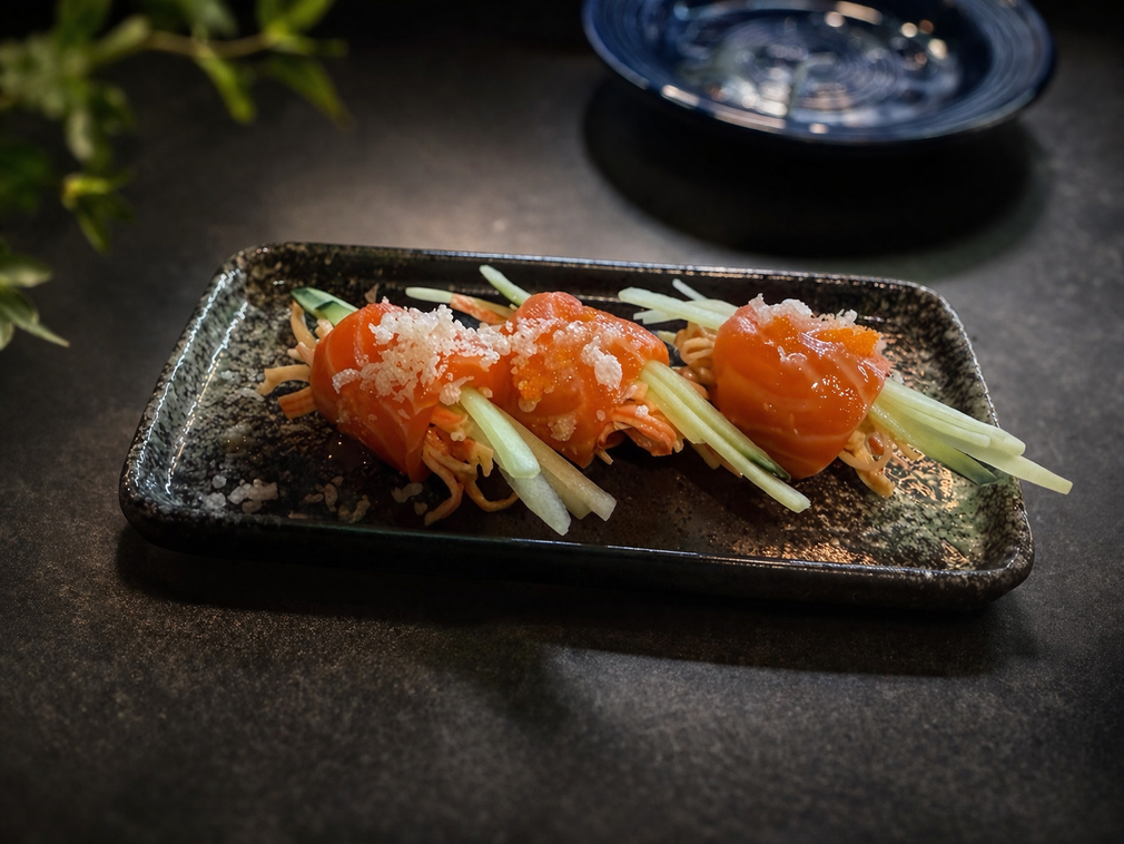 Three salmon rolls garnished with shredded cucumber on a dark, textured rectangular plate against a dim background.