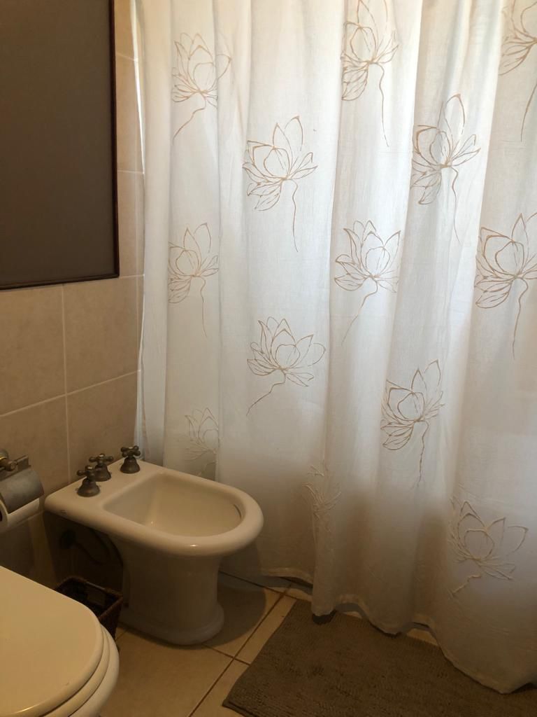Baño con inodoro blanco, bidé y cortina de ducha con estampado floral. También se aprecian paredes de azulejos beige y una pequeña alfombra.