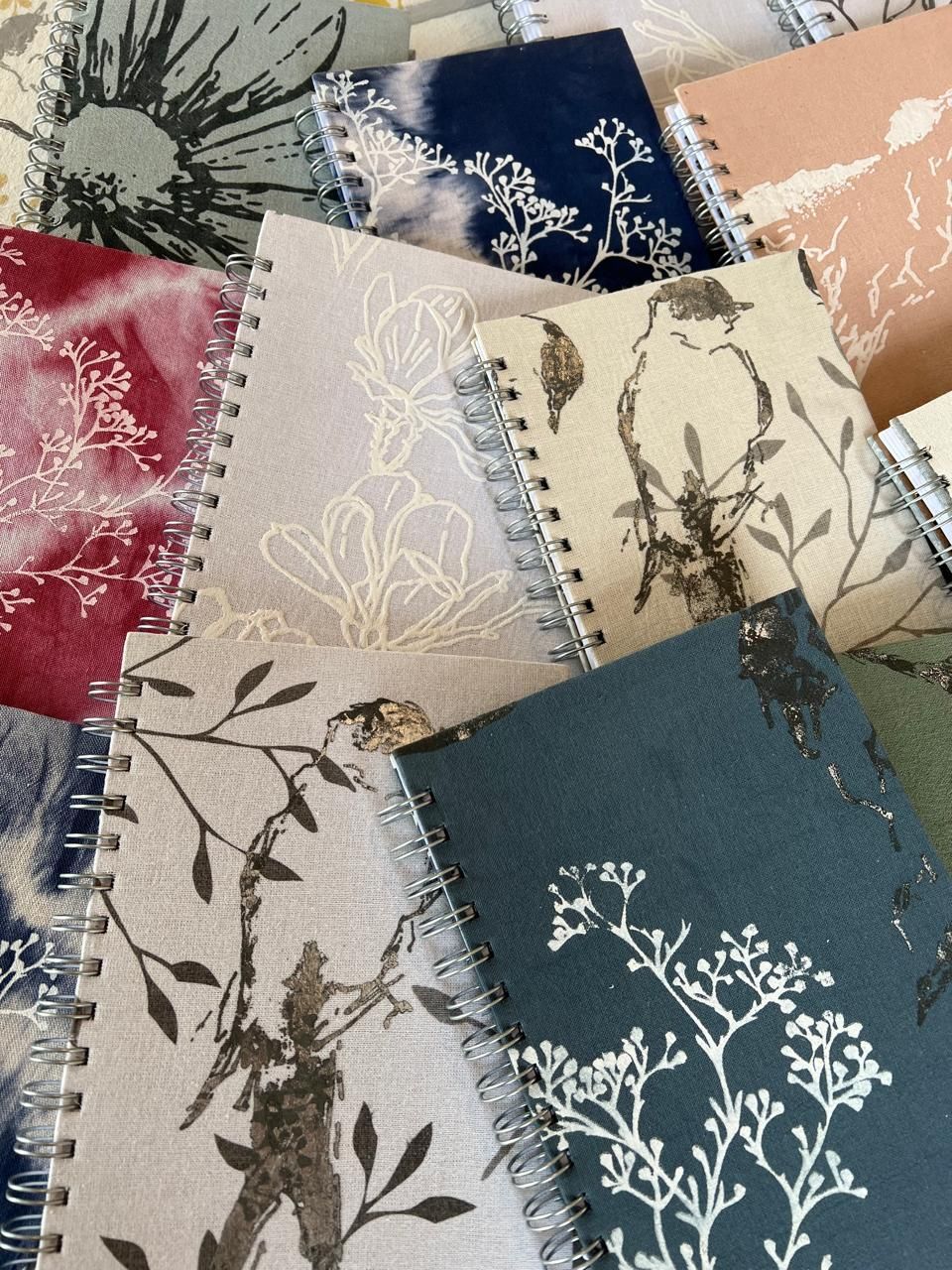 Surtido de cuadernos encuadernados en espiral con diseños florales y de pájaros sobre tapas de tela en varios colores.