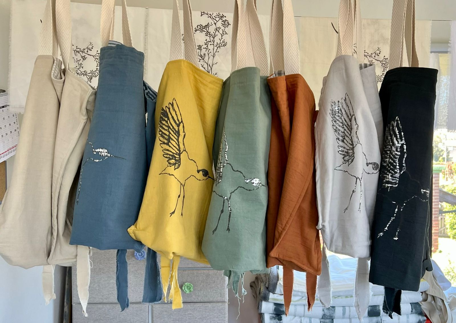 Siete bolsas de colores con largas correas blancas colgadas de ganchos sobre un fondo de madera desgastada.