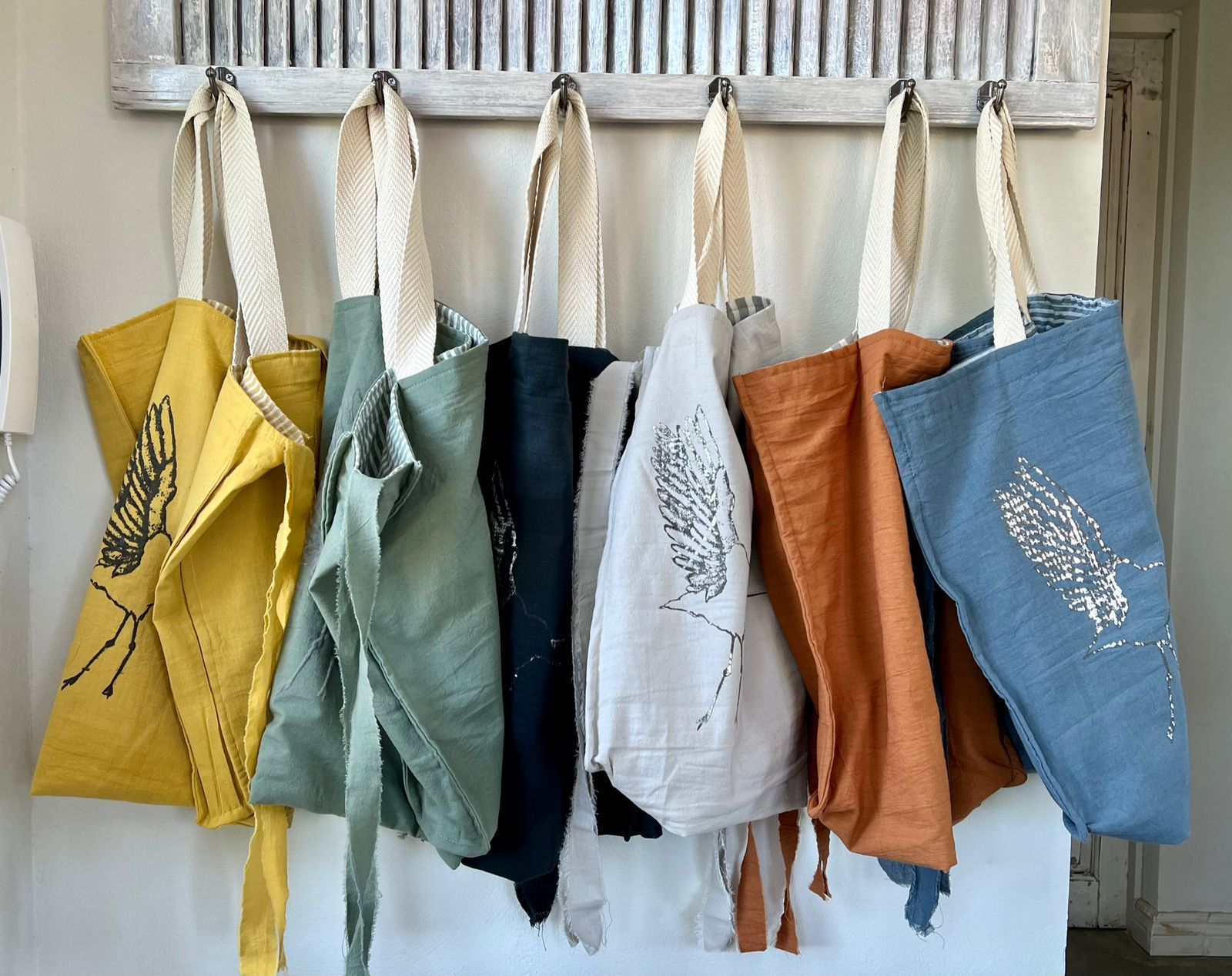 Siete bolsas de colores con largas correas blancas colgadas de ganchos sobre un fondo de madera desgastada.