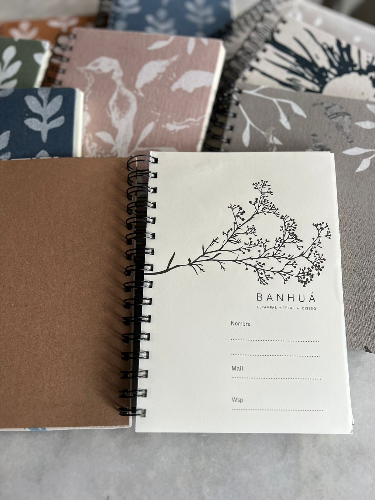 Una selección de cuadernos de espiral con estampado floral, uno abierto en una página con la etiqueta "MANHUA". Los cuadernos tienen una superficie clara.