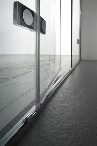 Porta scorrevole in vetro, con binario inferiore e maniglia argentata, che riflette il pavimento grigio e la parete bianca.