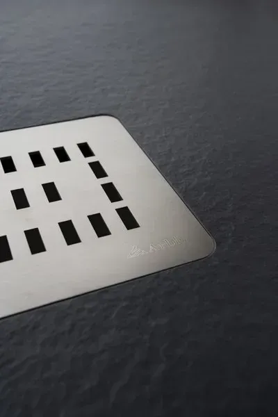 Piatto doccia in metallo quadrato con aperture rettangolari, inserito in un pavimento grigio scuro.