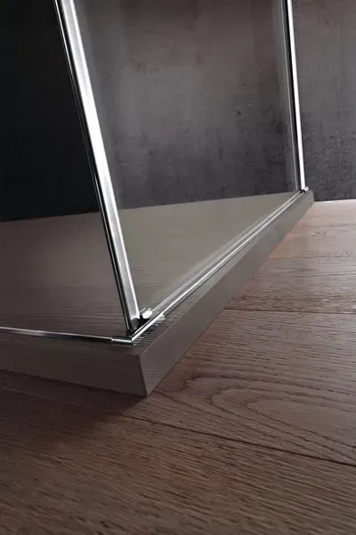 Primo piano dell'angolo di un box doccia con bordi metallici, appoggiato su un pavimento in legno.