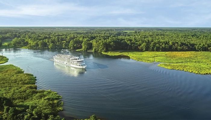 An aerial view of a cruise ship sailing down a river surrounded by trees.