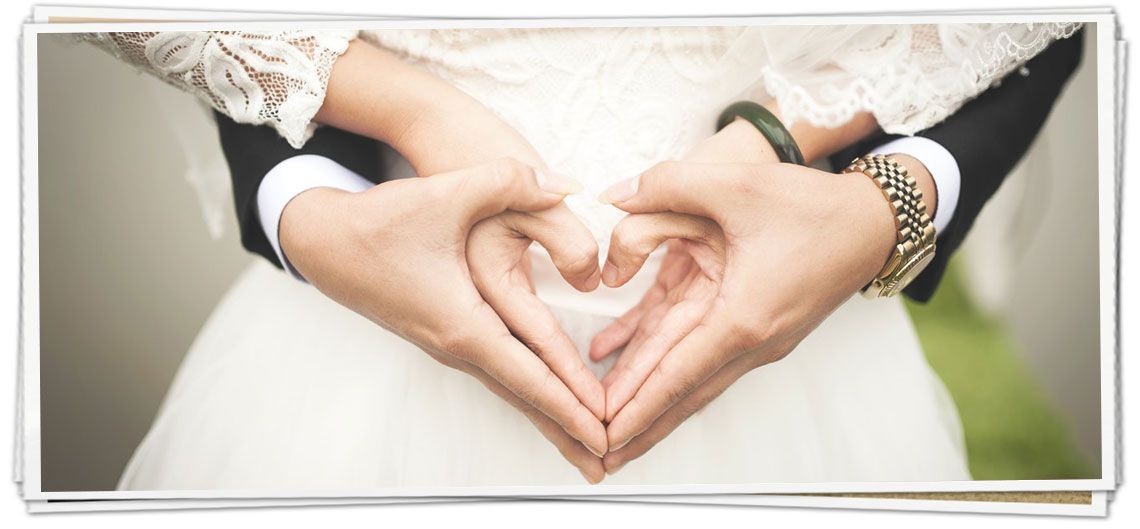 A bride and groom are making a heart shape with their hands.