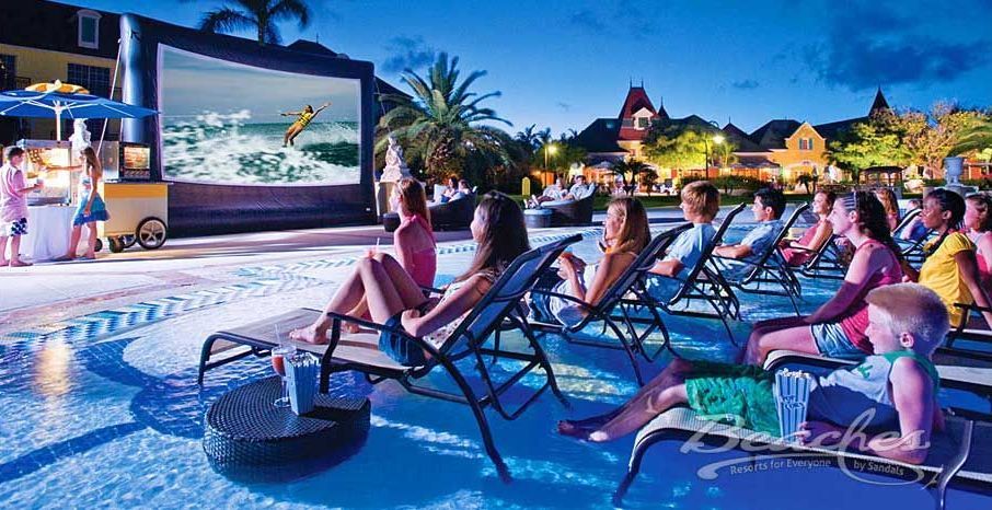 A group of people are sitting around a swimming pool watching a movie on a large screen.