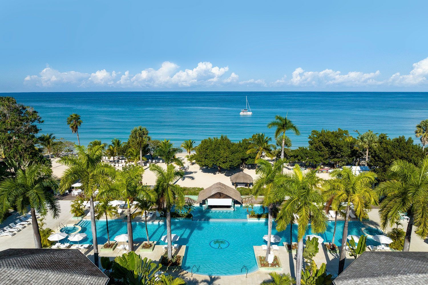 An aerial view of a resort with a large swimming pool surrounded by palm trees and a beach.