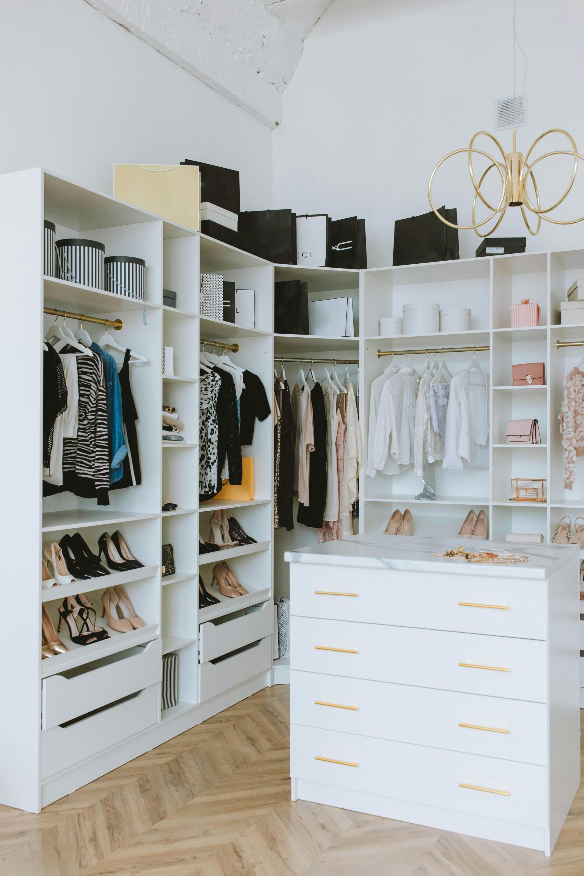 Bright walk-in closet with white shelves, hanging clothes, shoes, and drawers, lit by a gold chandelier