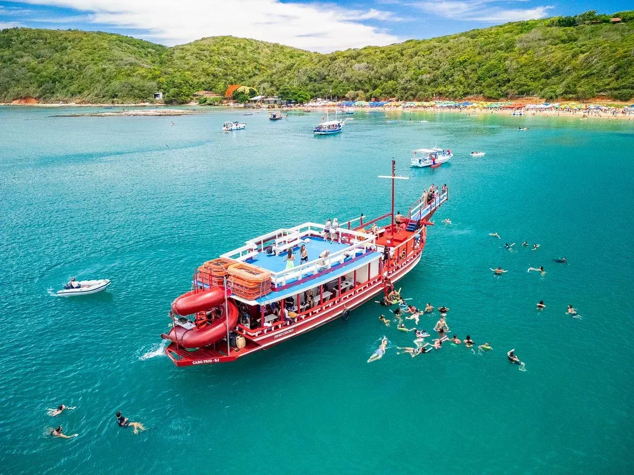Un gran barco rojo repleto de gente navegaba en aguas turquesas cerca de una playa y colinas verdes.