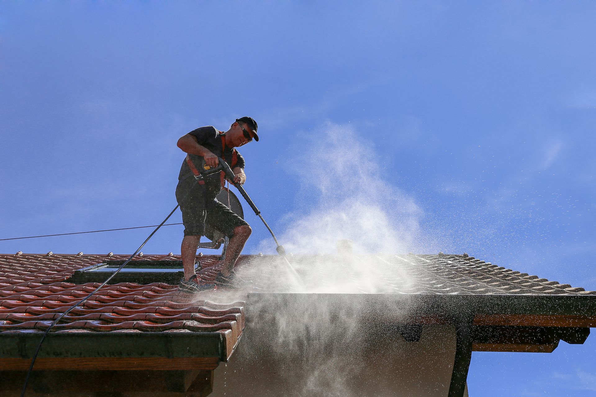 Worker power washing a red-tiled roof with water spraying into the air under a blue sky