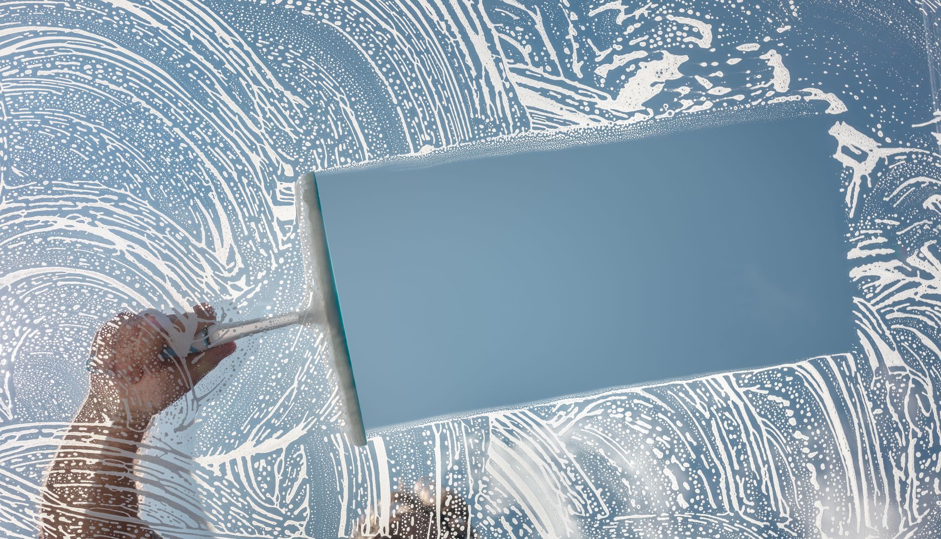 Hand holding a squeegee on a frosted glass window, clearing a blue-tinted area.