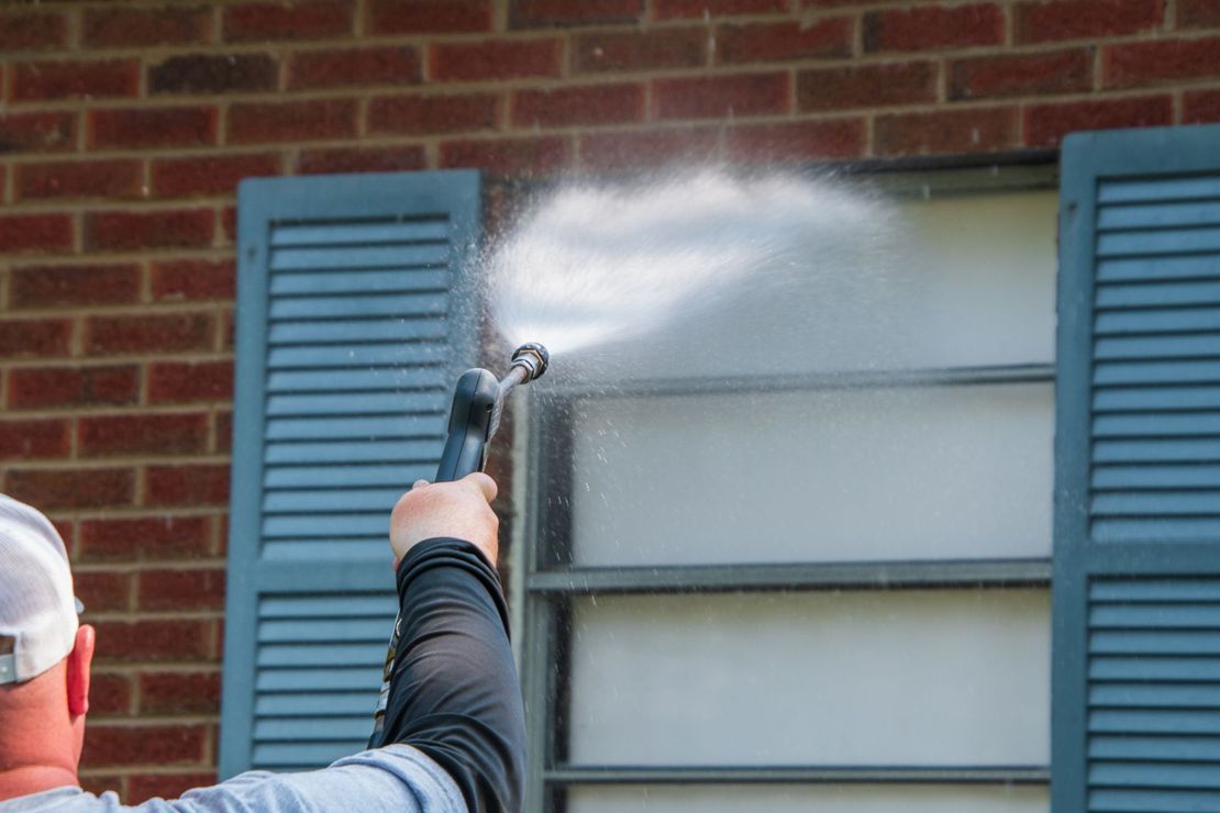 Person pressure-washing a blue shuttered window on a brick house
