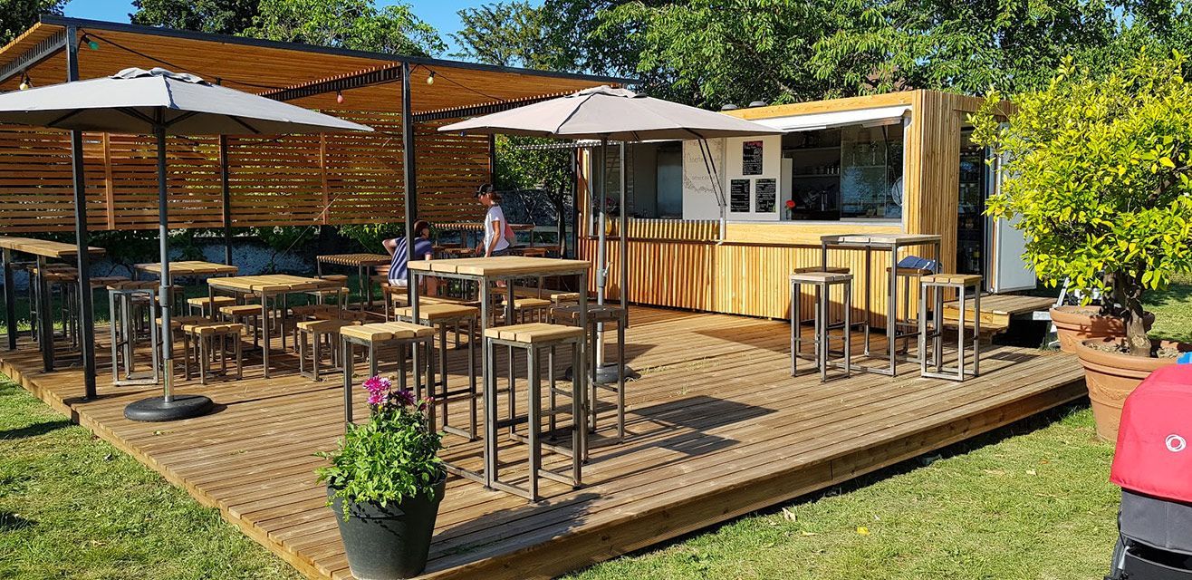 Restaurant en plein air avec terrasse, tables, parasols et bar à conteneurs, installé sur l'herbe sous un ciel bleu.