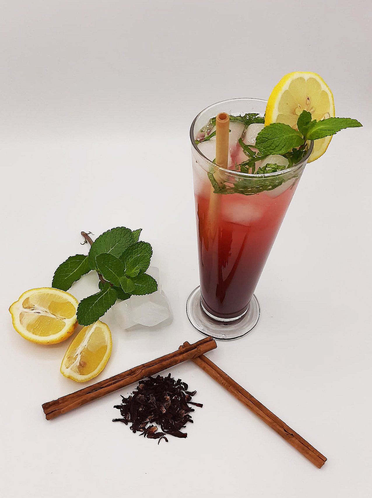 Une boisson rouge vif dans un grand verre, garnie de citron et de menthe, à côté d'ingrédients comme des bâtons de cannelle et du thé en vrac.