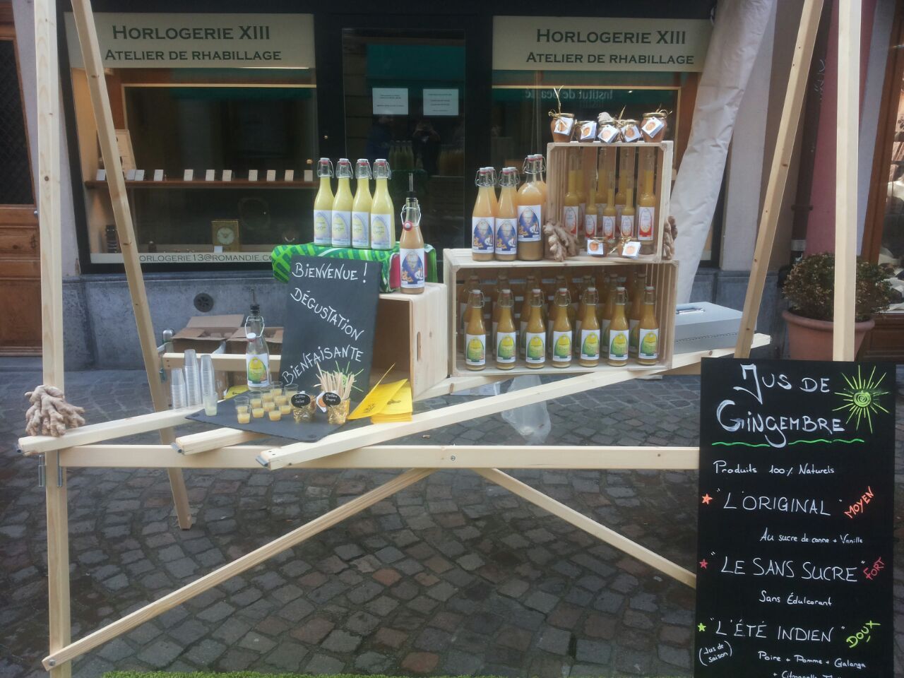 Stand de jus de gingembre dans la rue, bouteilles exposées sur une structure en bois. Menu à l'ardoise noire.