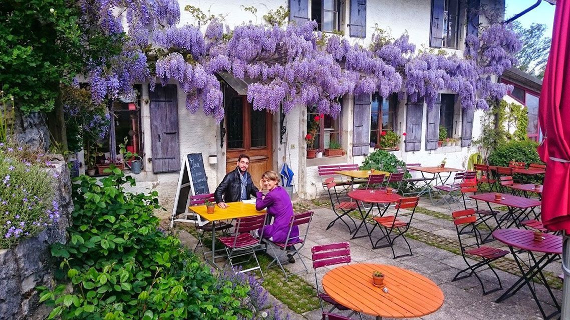 Café avec glycine violette, deux personnes à une table. Tables et chaises rouges et orange sur une terrasse en pierre.