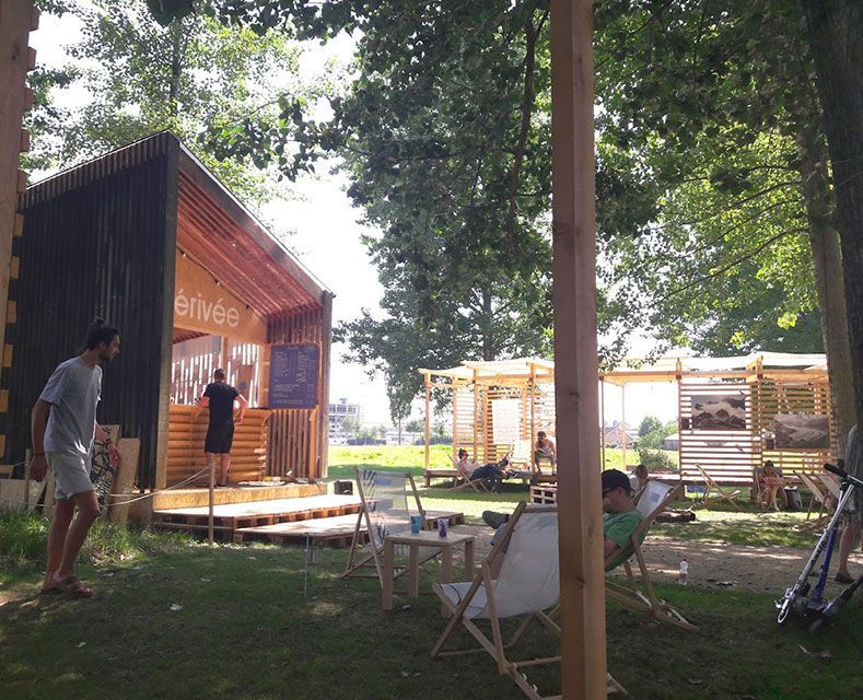 Café en plein air avec des structures en bois et des personnes se relaxant sur des chaises, des arbres en arrière-plan.