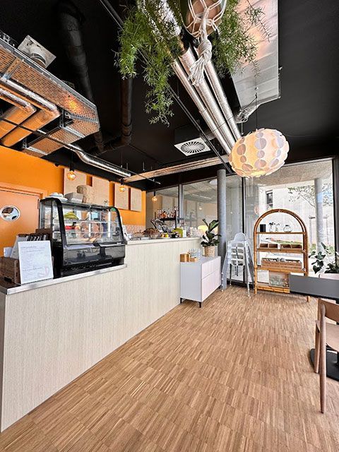 Intérieur d'un café avec parquet, comptoir blanc et plantes suspendues. Plafond noir, mur orange et grande fenêtre.