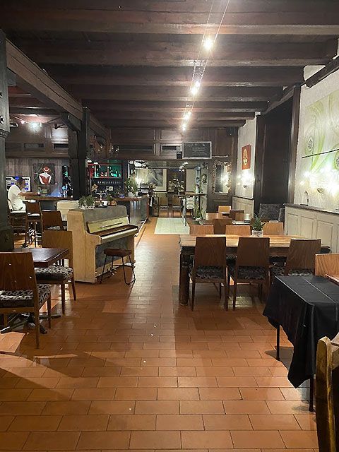 Vue intérieure d'un restaurant avec poutres en bois, tables, chaises, piano et coin bar.
