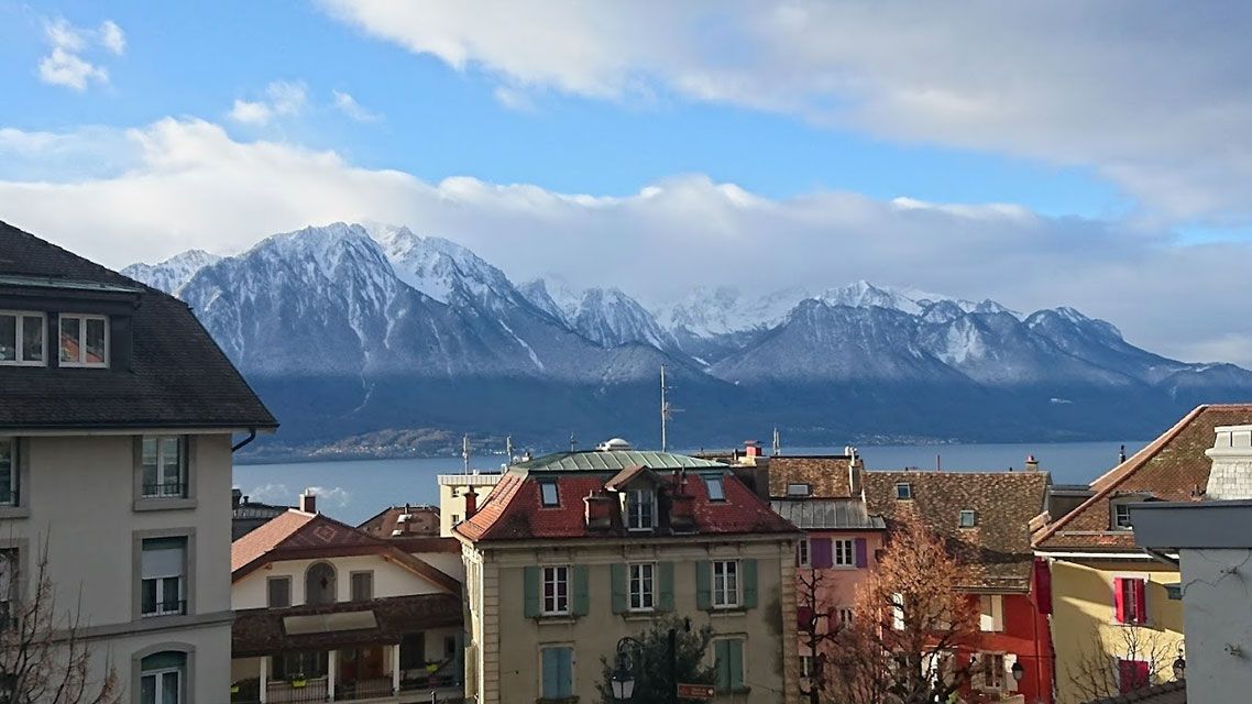 Les bâtiments surplombent un lac et des montagnes enneigées sous un ciel bleu vif avec des nuages.
