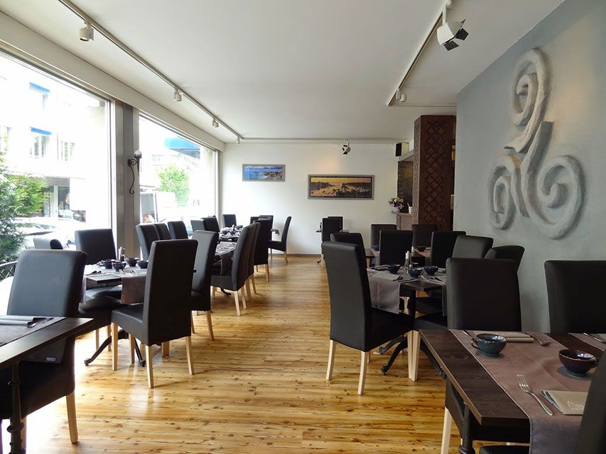 Intérieur de restaurant avec plancher en bois, tables dressées pour dîner, grandes fenêtres et une sculpture murale décorative.
