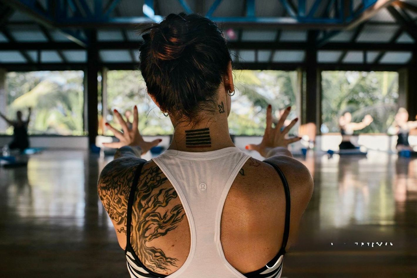 Person with tattooed back raises hands in a yoga studio with mirrors and large windows.