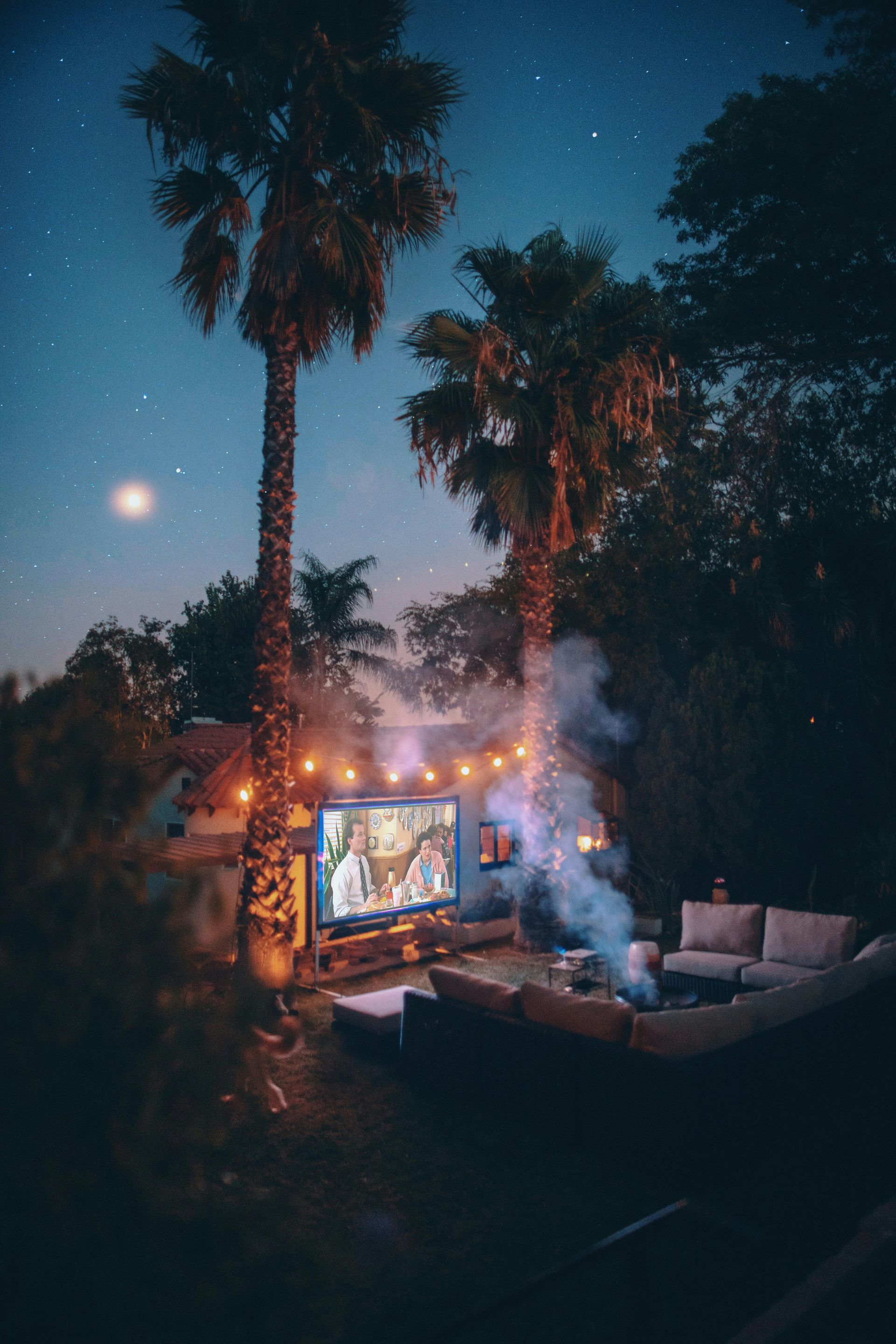 Outdoor movie setup with a glowing screen, string lights, and smoke among palm trees at dusk