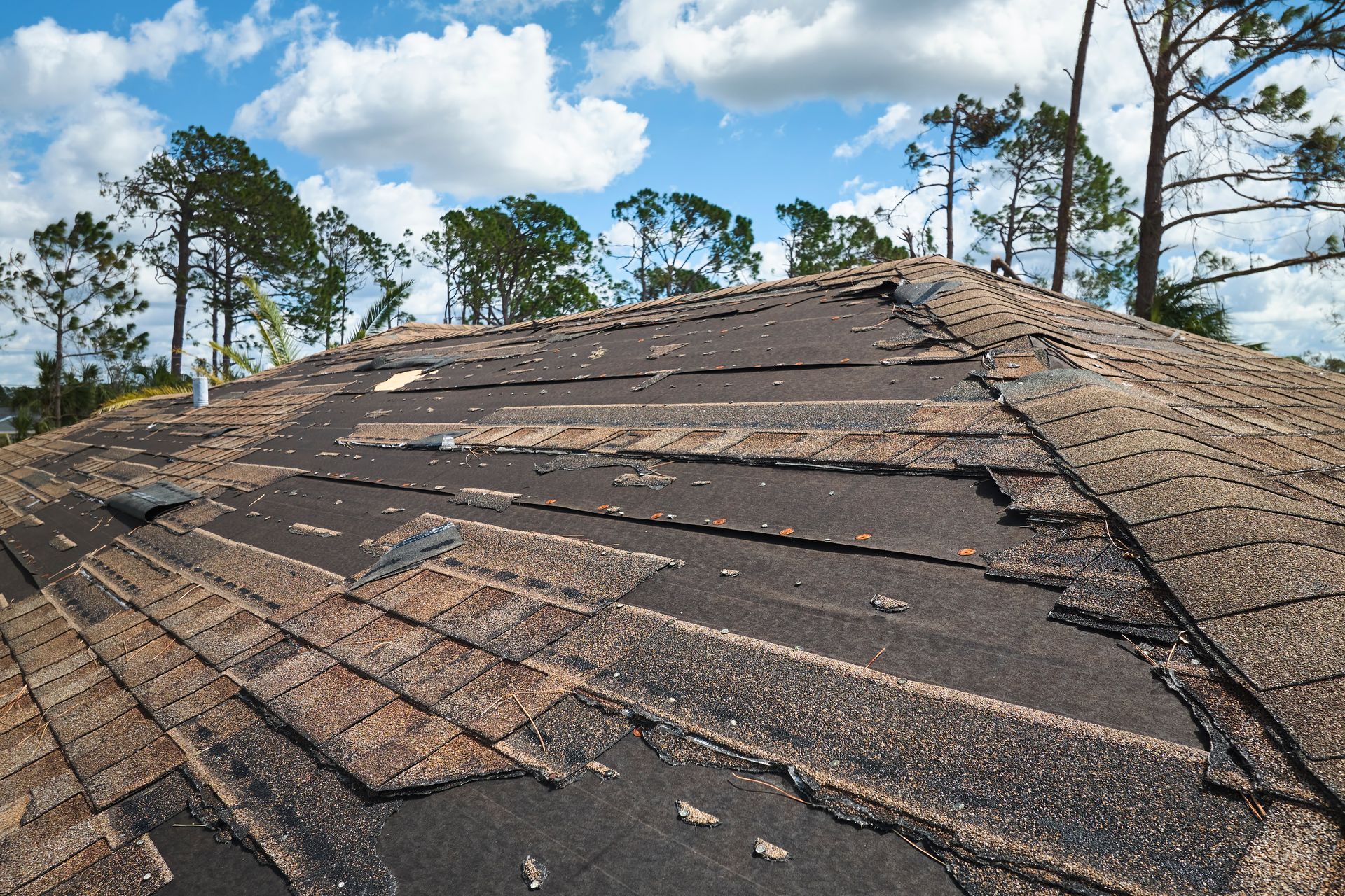 Damaged asphalt shingle roof, with missing and worn shingles, against a cloudy sky.