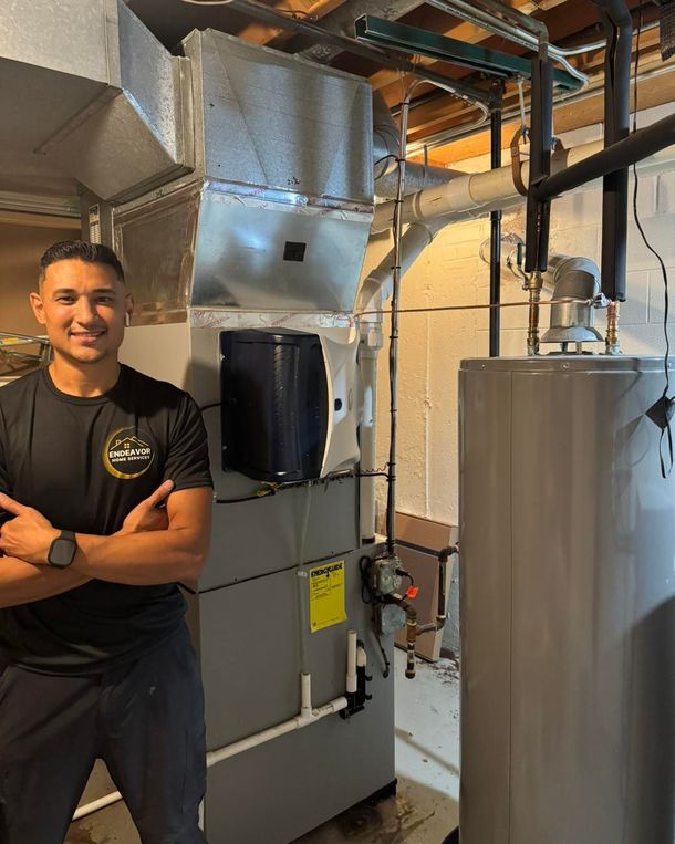 Man in black shirt stands next to HVAC unit and water heater in a utility room.
