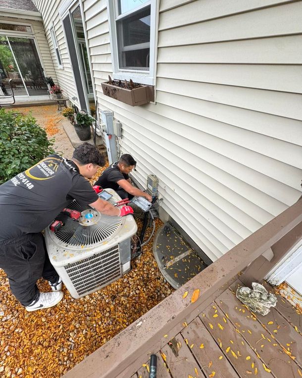 Two people installing an air conditioning unit near a house's wall. Fallen leaves cover the ground.
