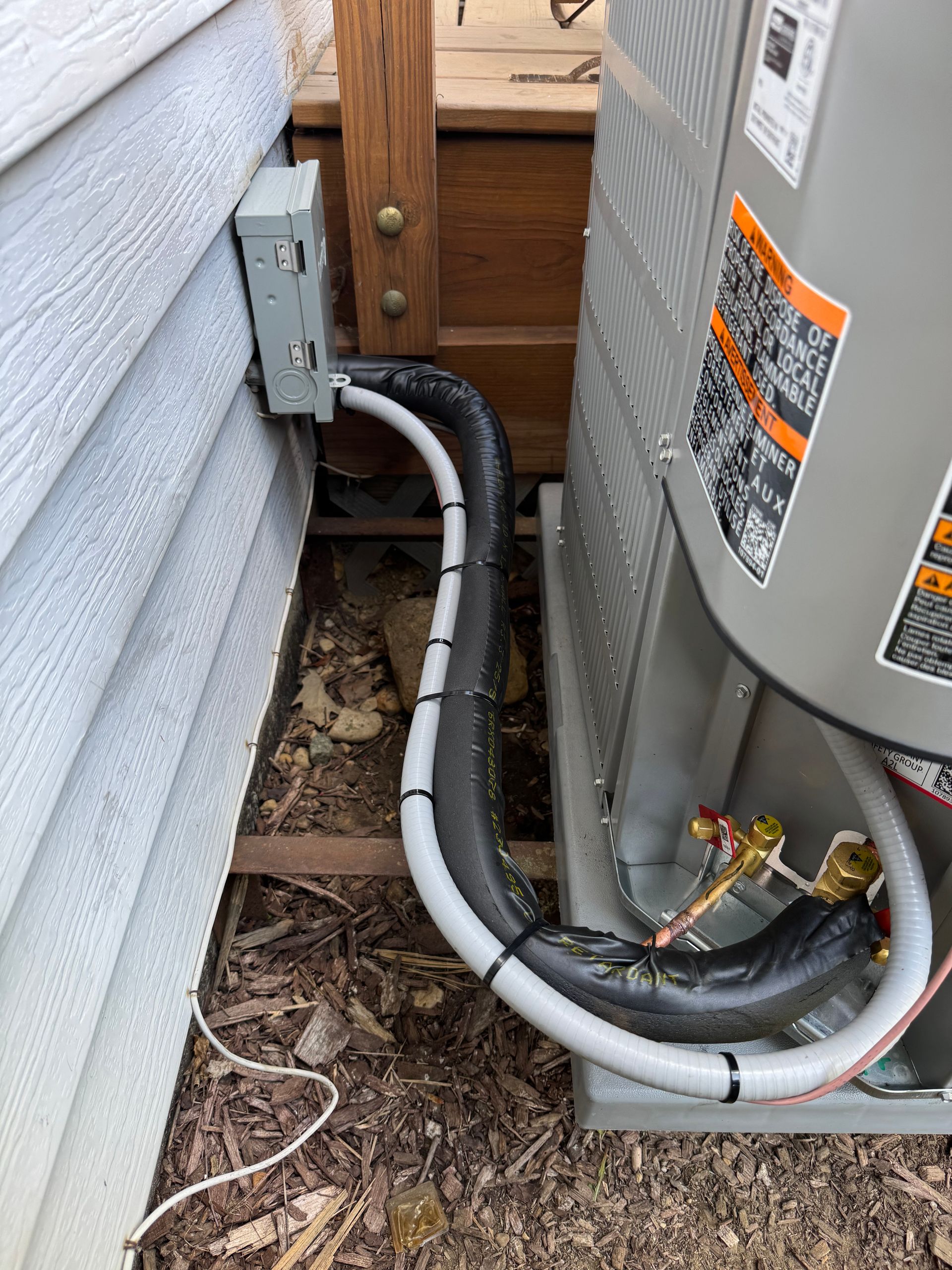 Air conditioner unit next to a house with insulated pipes. Electrical box visible on the wall.